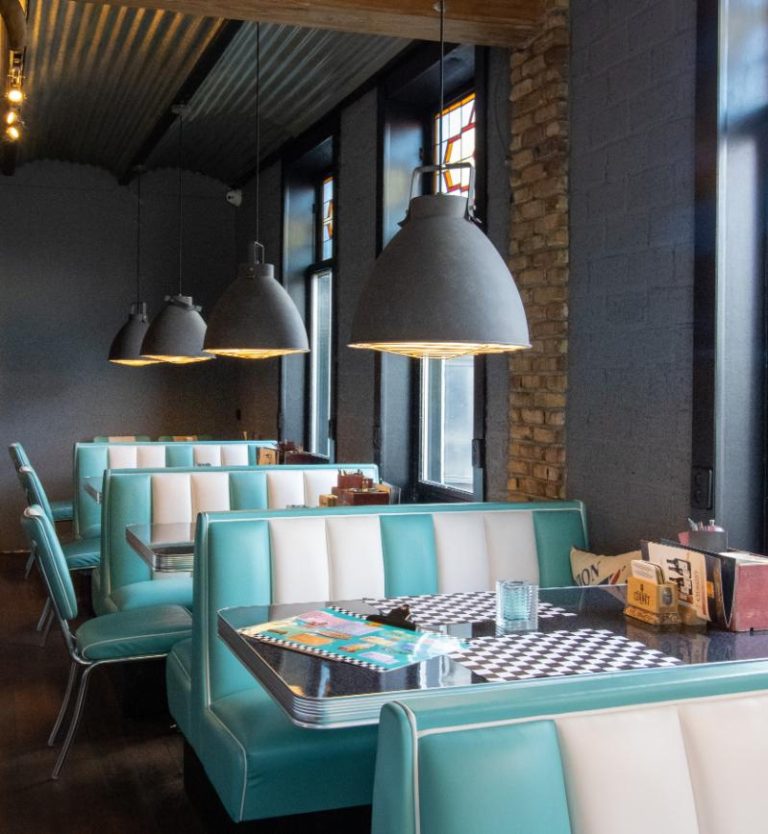 Retro diner met blauwgroene en witte banken, dambordtafels en grijze industriële lampen.
