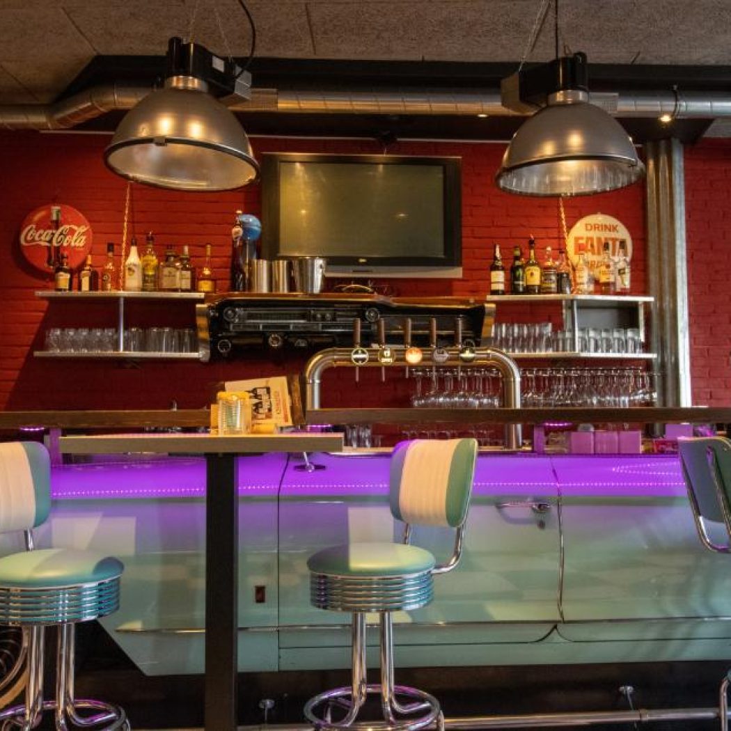 Bar in Amerikaanse retrostijl met een lichtblauwe oldtimer als bar, paarse verlichting en chromen krukken.