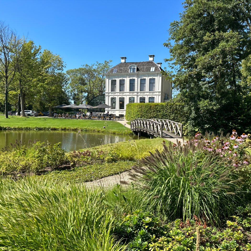 Wit landhuis met buitenterras aan een vijver met houten bruggetje, omgeven door groene tuinen.