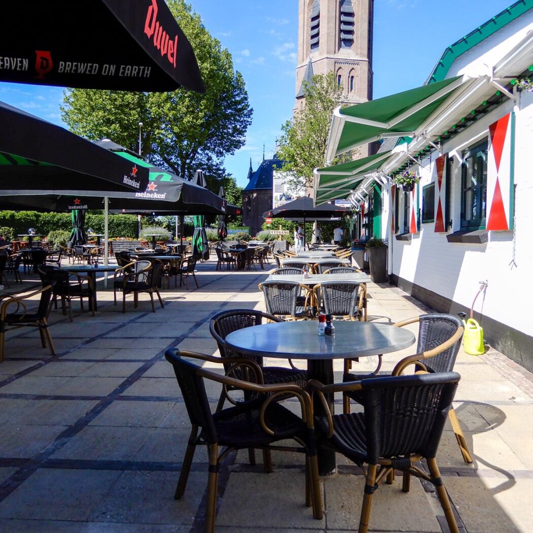 Zonnig caféterras met Duvel parasols (Tastes like Heaven), wit gebouw met rood-wit gestreepte luiken en kerktoren.