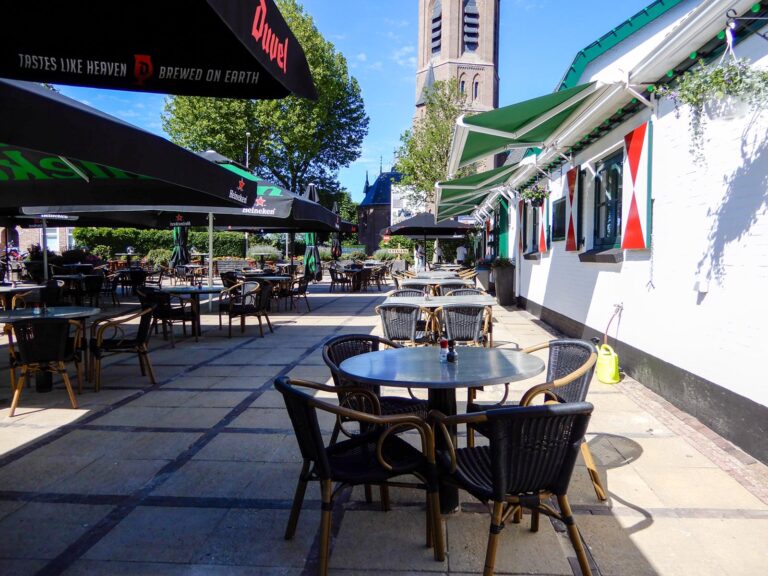Zonnig caféterras met Duvel parasols (Tastes like Heaven), wit gebouw met rood-wit gestreepte luiken en kerktoren.