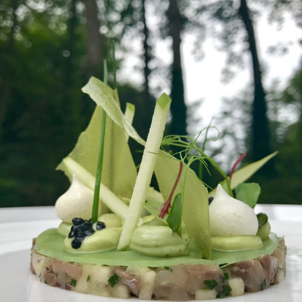 Verfijnd groen voorgerecht met fijngehakte basis, verse groene garnering, witte mousse en kaviaar, tegen een wazige bosachtergrond.