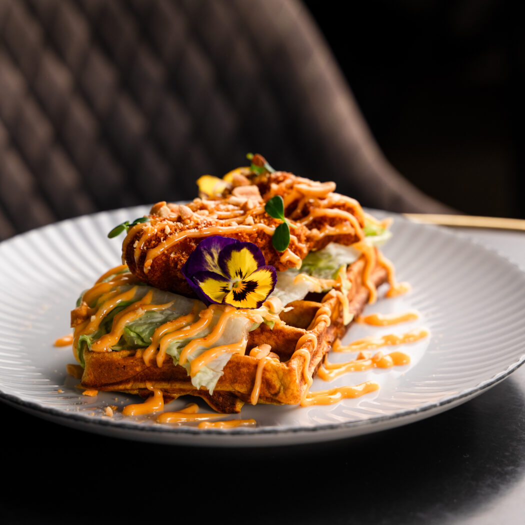 Krokante kip op een wafel met verse sla, oranje saus, pindas en een paarse eetbare bloem op wit bord.
