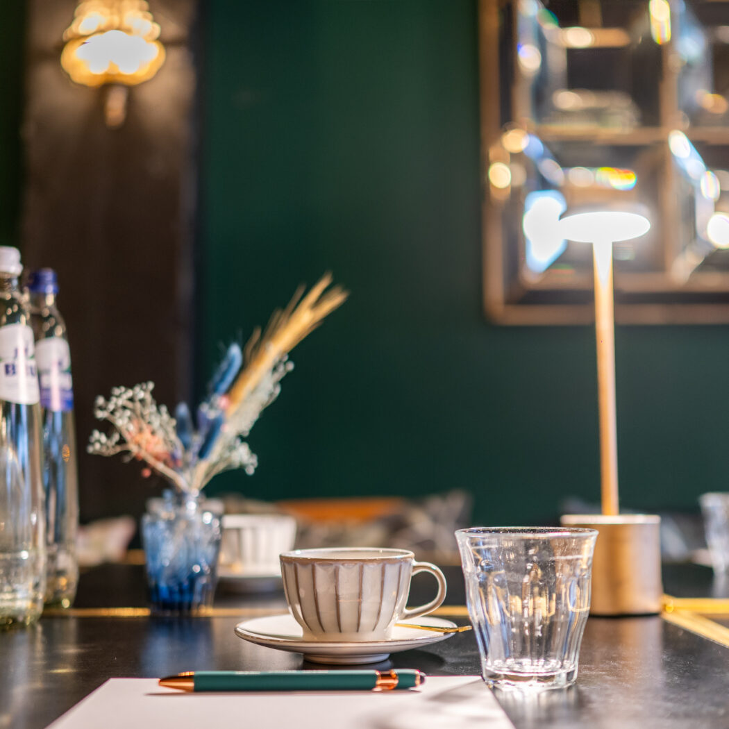 Gedekte tafel met J. BLANC waterflessen, gestreepte koffiekop, waterglas, pen en tafellamp tegen groene muur.