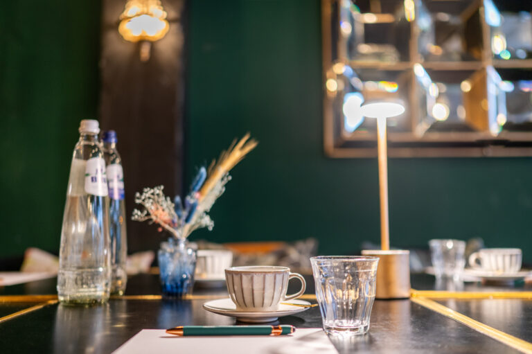 Gedekte tafel met J. BLANC waterflessen, gestreepte koffiekop, waterglas, pen en tafellamp tegen groene muur.
