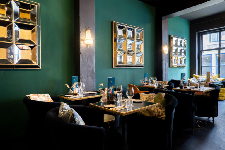Sfeervol restaurant met diepgroene muren, fluwelen stoelen, gedekte tafels en decoratieve spiegels.