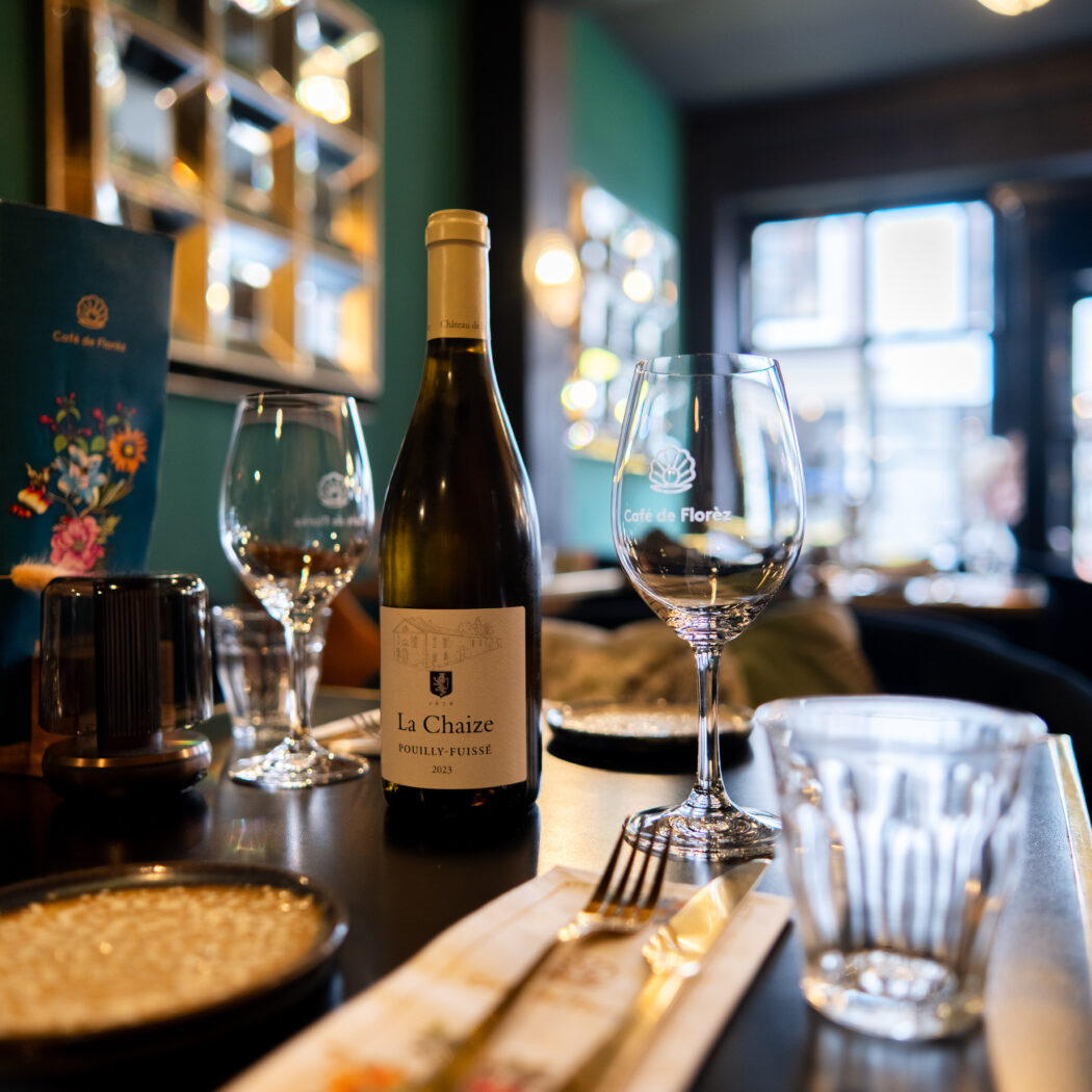 Fles La Chaize Pouilly-Fuissé witte wijn, wijnglazen met Café de Florez logo, menu en gedroogde bloemen op tafel.