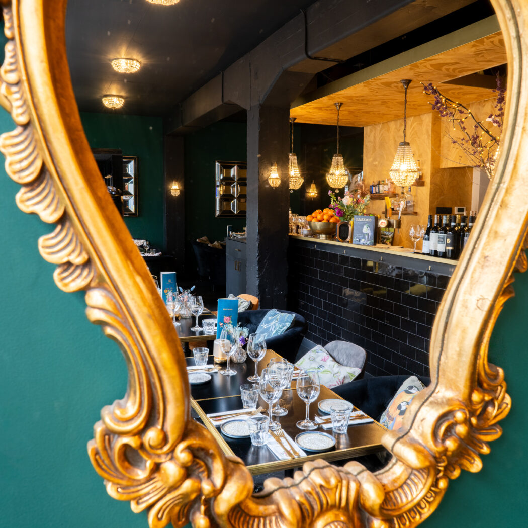 Goudkleurige, sierlijke spiegel aan een donkergroene muur, weerspiegelt een chic restaurant met gedekte tafels en bar.