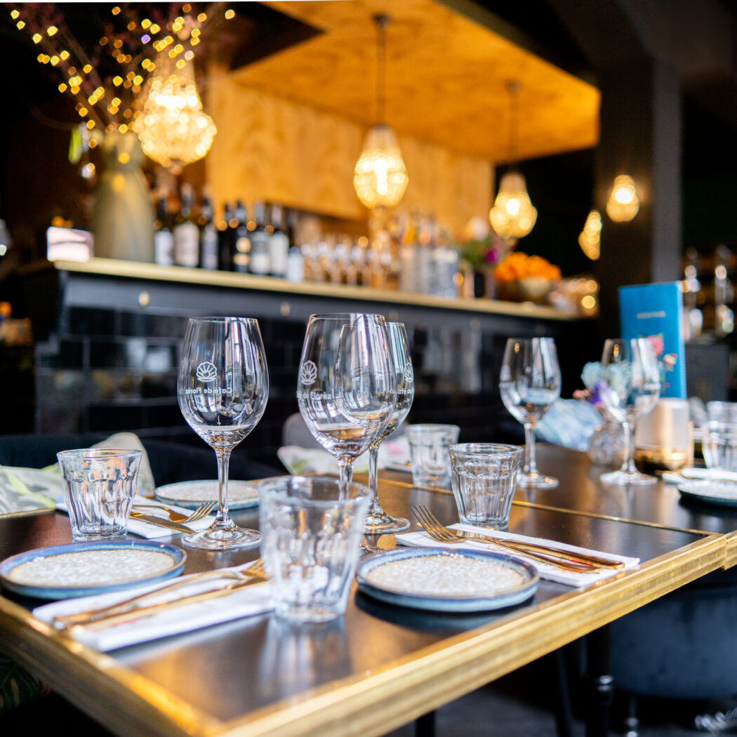 Gedekte restauranttafel met wijnglazen met logo, borden en goudkleurig bestek in een sfeervol interieur.
