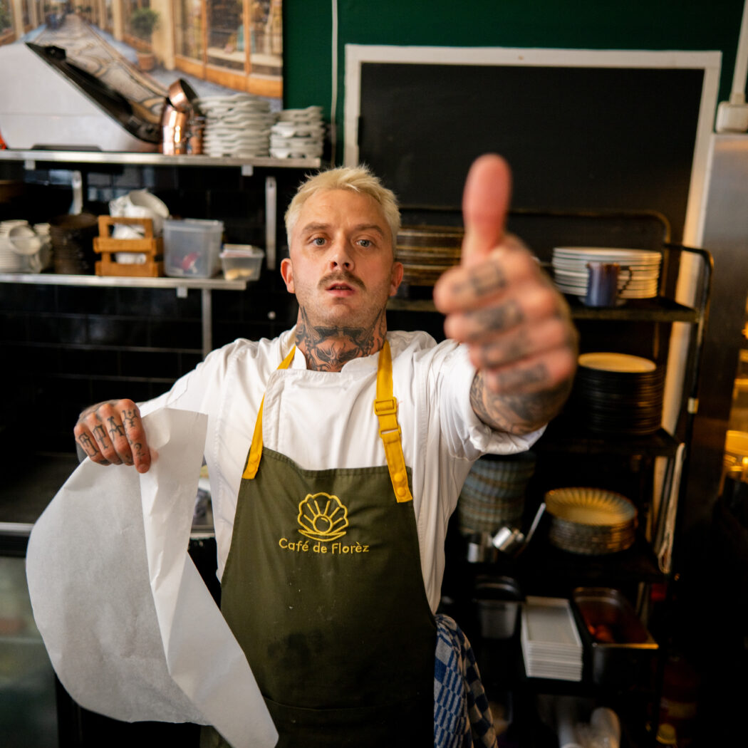 Getatoeëerde chef in Café de Florez schort geeft een duim omhoog en houdt bakpapier vast in de keuken.