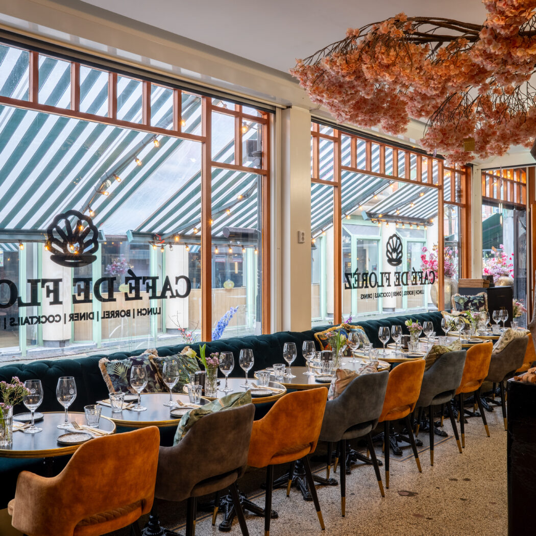 Interieur van Cafe De Flores met gedekte tafels, groene bank, oranje stoelen en bloesemboom bij grote ramen.