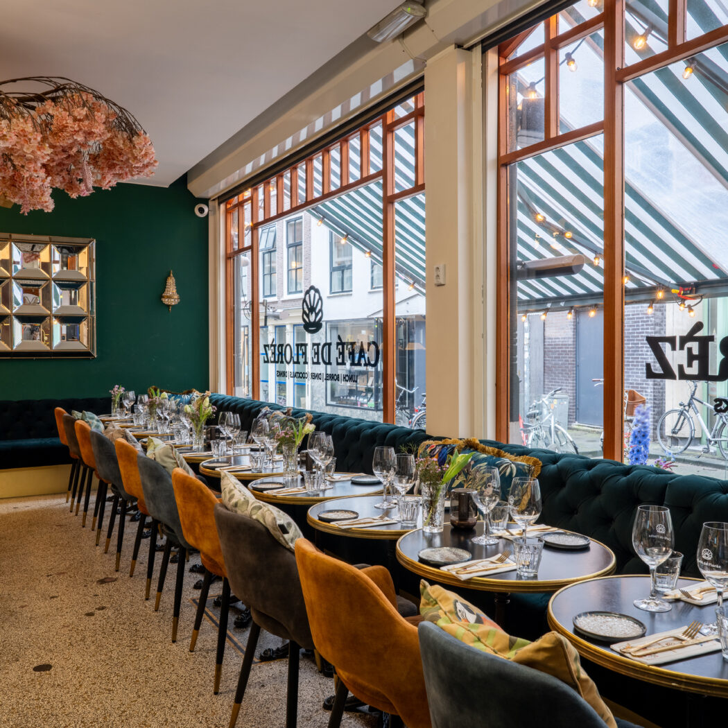 Donkergroen restaurantinterieur met fluwelen bank, ronde tafels, kersenbloesemboom. CAFE DE FLORES op het raam.