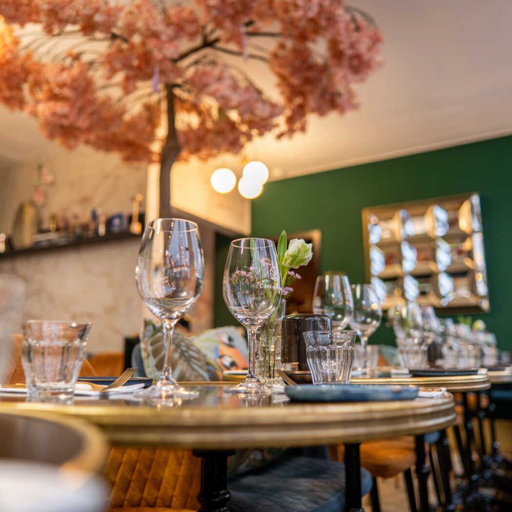 Gedekte tafels in een sfeervol restaurant met gouden accenten, groene muren en roze bloesem decoratie aan het plafond.