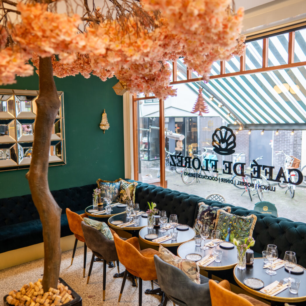 Gezellig interieur van Cafe de Flores met bloesemboom, teal bank, sierkussens en gedekte tafels.