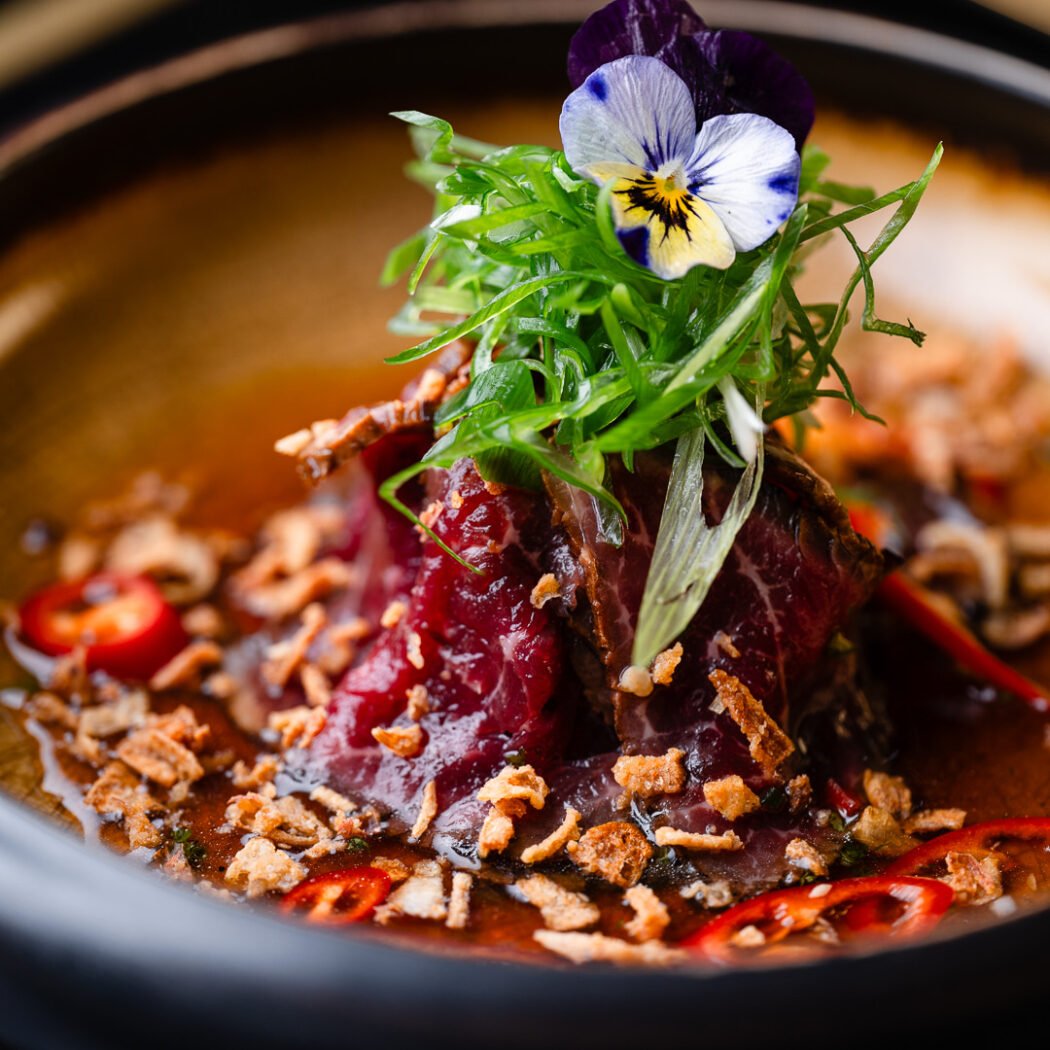 Luxe gerecht met dun gesneden rood vlees, frisse groene kruiden, krokante uitjes, rode peper en een paars-wit viooltje.