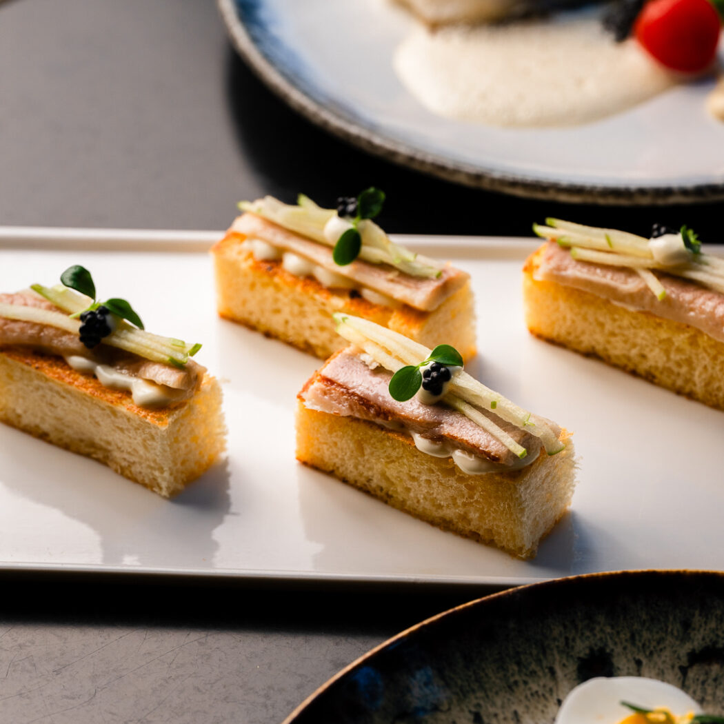 Vier canapés op geroosterd brood met fijne beleg, groene appel en kaviaar op een witte schaal.