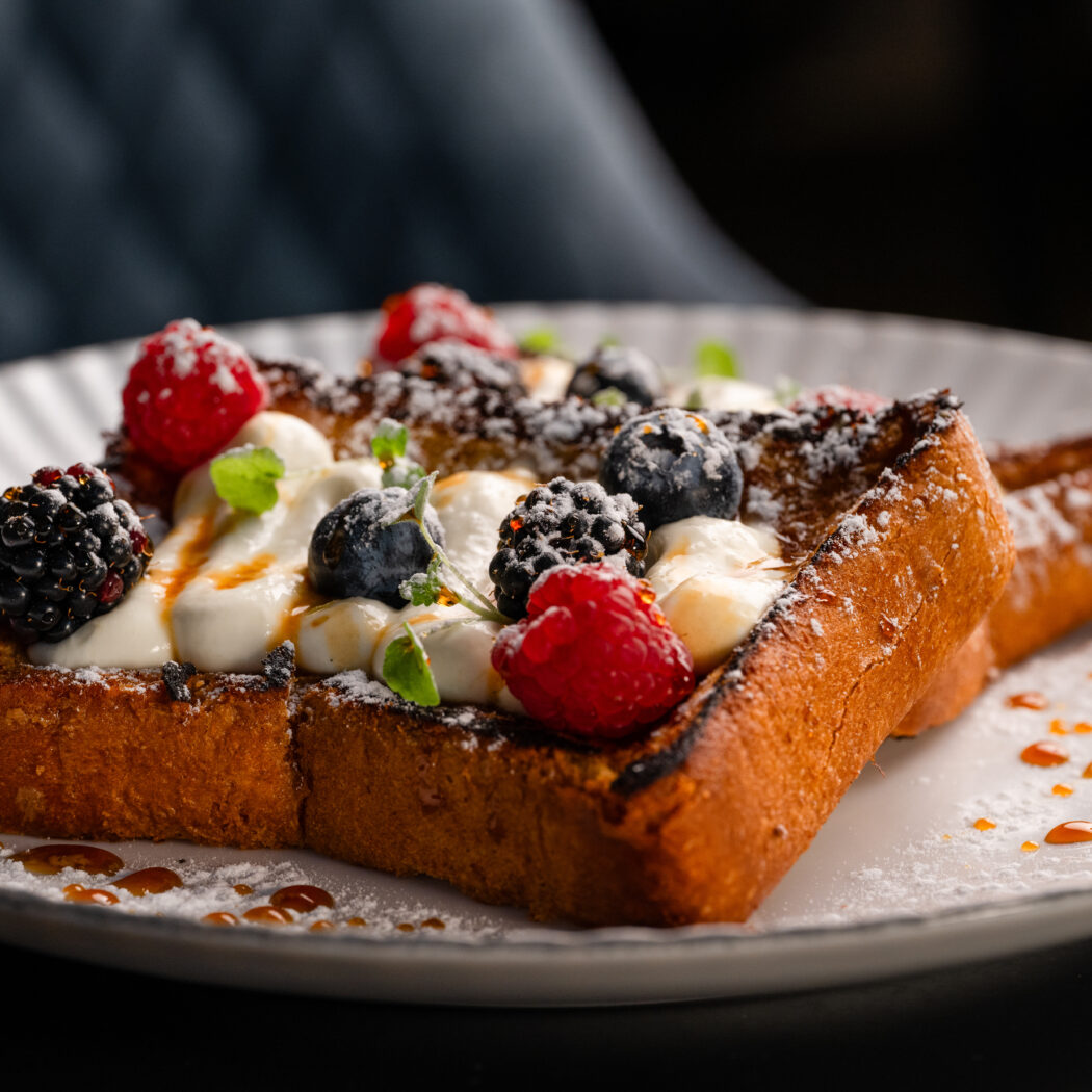 Wentelteefjes met diverse bessen, slagroom, poedersuiker en een drizzle karamelsaus op een wit bord.