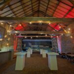 Sfeervolle evenementenlocatie met bakstenen muren, houten balken, rode verlichting en statafels.