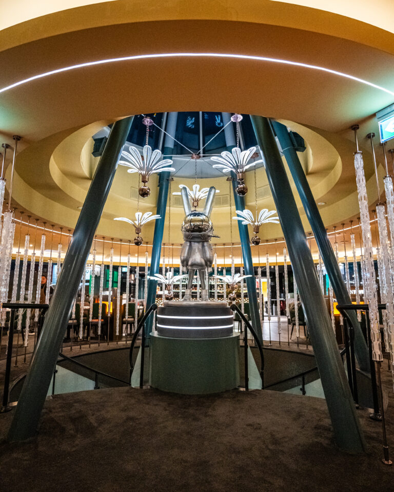 Grote zilveren konijnsculptuur op verlicht voetstuk, omringd door sierlijke hanglampen in een modern interieur.
