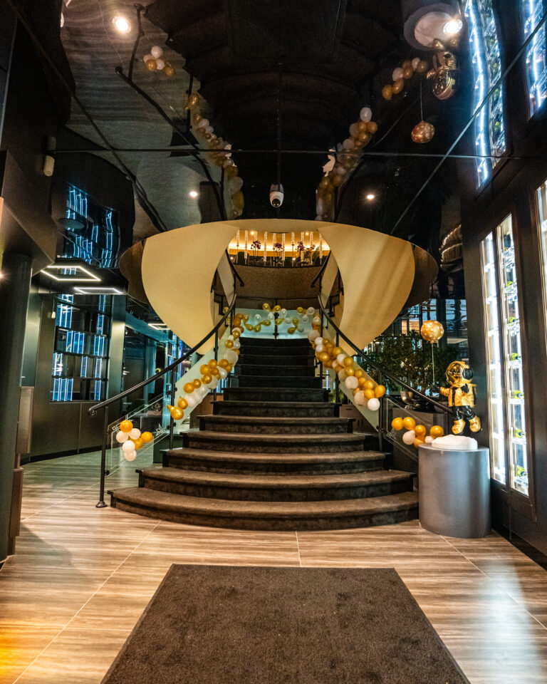 Een feestelijk versierde, gebogen trap met goud-witte ballonnen en een gouden boog in een modern interieur.