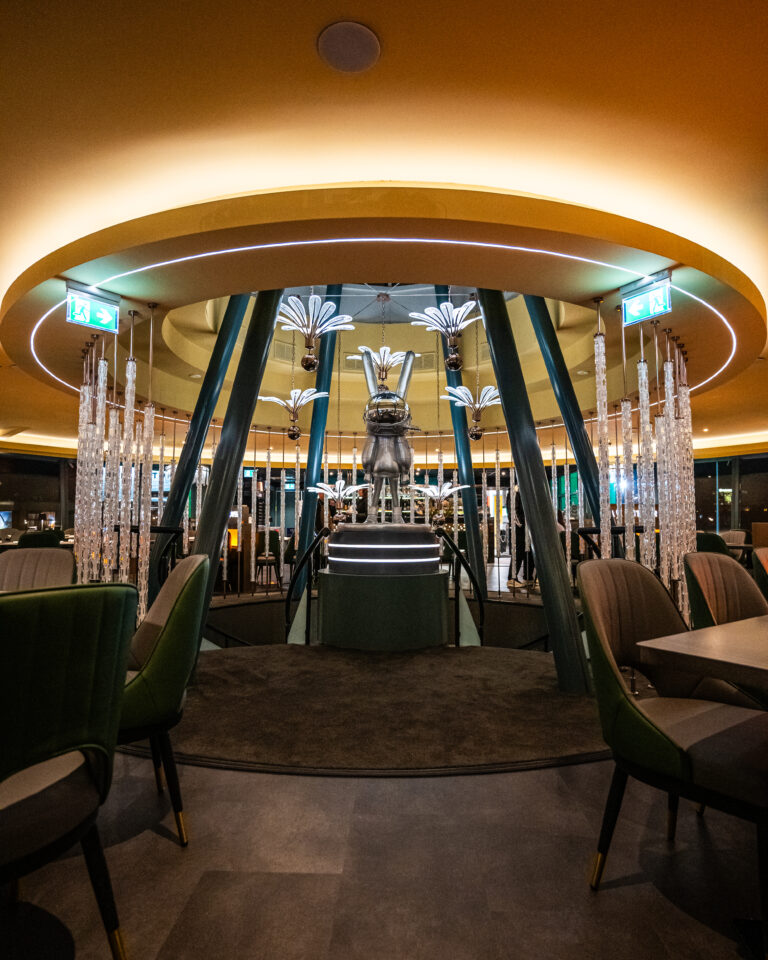 Glimmende konijn sculptuur op verlicht podium in luxe ronde lounge met moderne kroonluchters en groene stoelen.