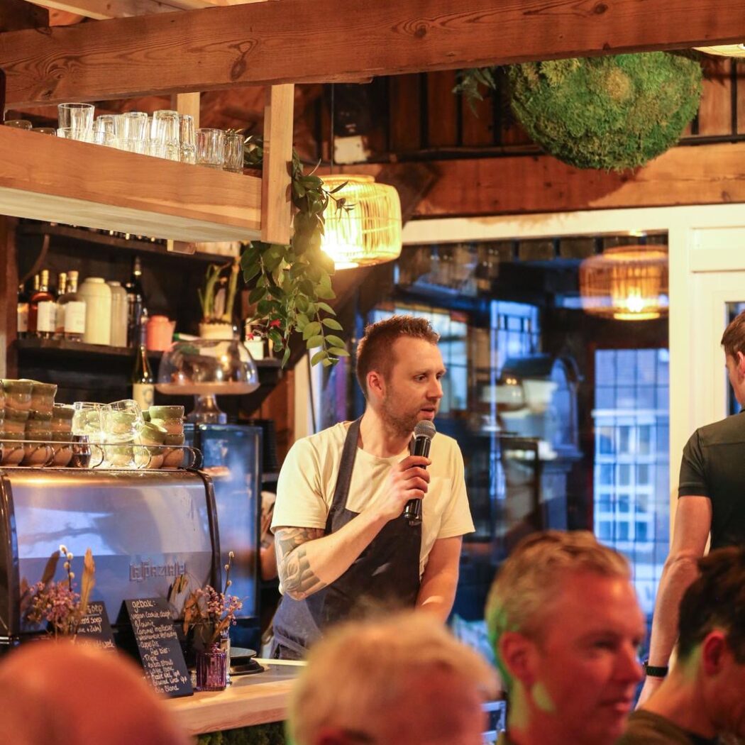 Man met schort spreekt in microfoon achter bar, met een bord Gebak en sfeervolle houten architectuur.