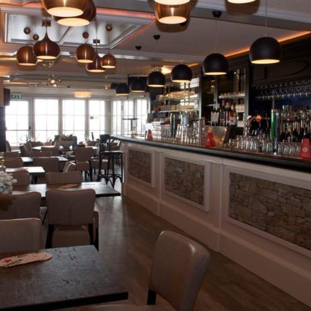 Warm verlicht restaurantinterieur met een lange, gevulde bar, diverse tafels, grote ramen en witte bloemen.