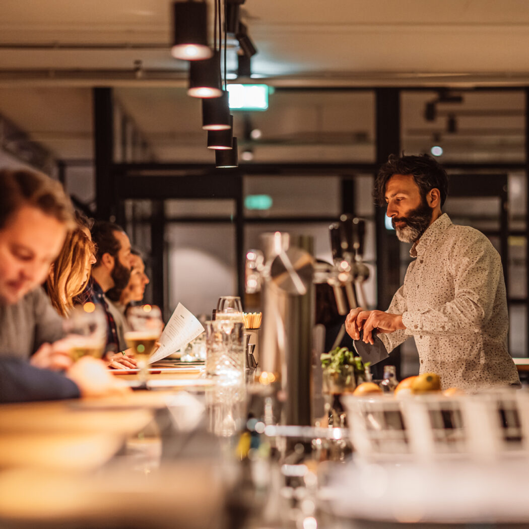 Een bebaarde barman met wit patroonoverhemd bereidt een drankje achter een moderne bar vol met klanten.