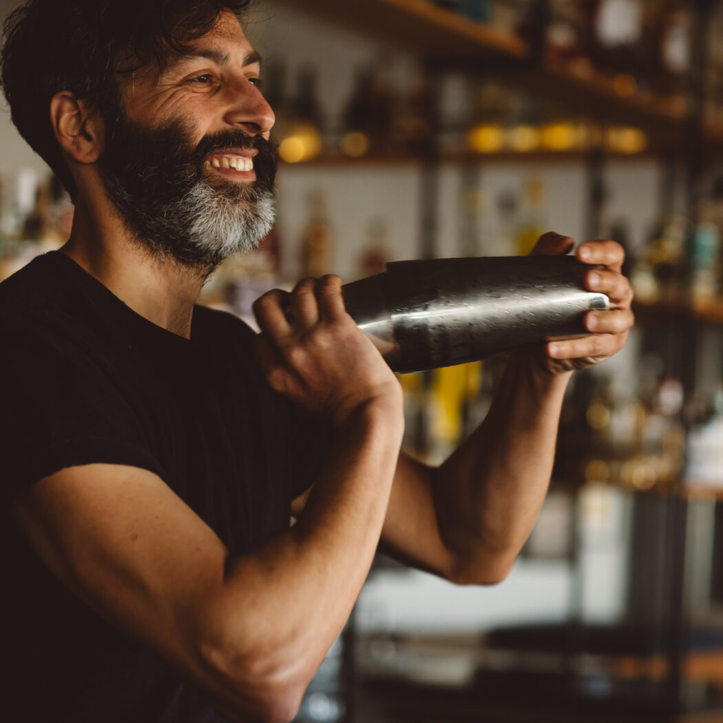 Lachende bartender met donkere baard die een zilveren cocktailshaker schudt in een bar.