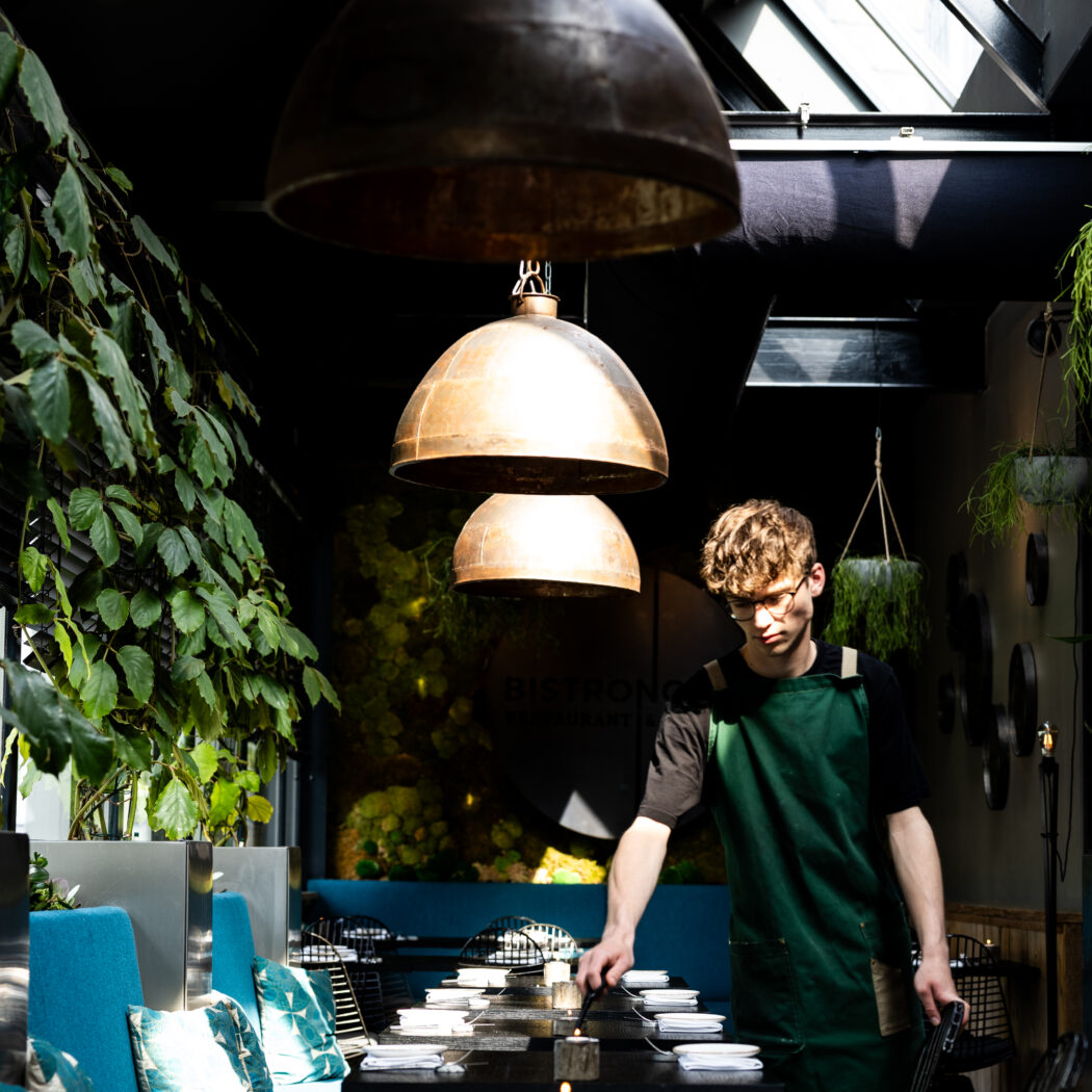 Jonge man in groen schort dekt tafels in een sfeervol restaurant met blauwe banken en grote koperen lampen.