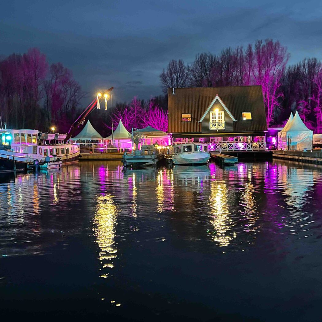Feestelijke waterlocatie met aangemeerde boten waaronder De Koningin, een pand en tenten, verlicht met roze en gele reflecties.