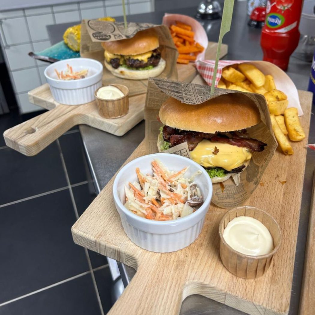 Twee burgers met kaas, spek, friet, coleslaw en mayonaise, geserveerd op houten snijplanken.