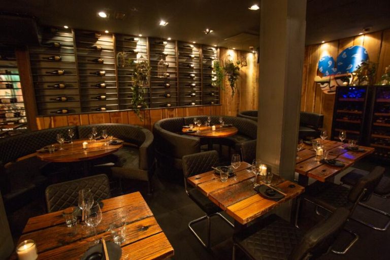Sfeervol restaurantinterieur met houten wanden, wijnrekken, donkere zitplaatsen en rustieke tafels met kaarslicht.