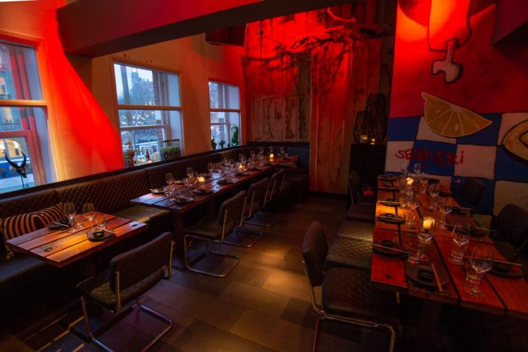 Sfeervol restaurantinterieur met lange houten tafels, donkere banken, rood licht en SENKORI op de muur.