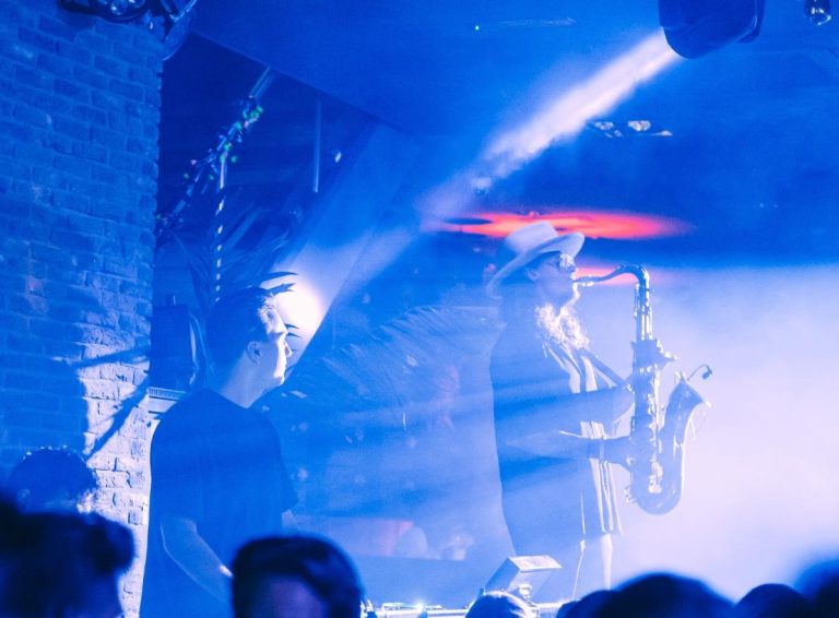 Saxofonist met hoed en zonnebril speelt op een podium onder felle blauwe lichten en rook.