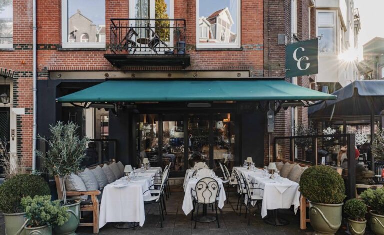 Uitnodigend restaurantterras van CG met groene luifel, gedekte witte tafels en planten voor een bakstenen gevel.