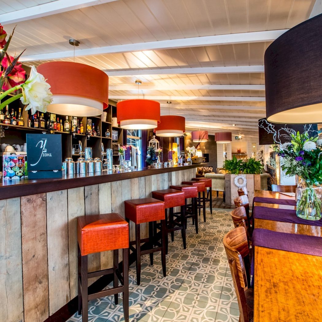 Stijlvol restaurantinterieur met een houten bar, oranje barkrukken, diverse bloemvazen en grote hanglampen.