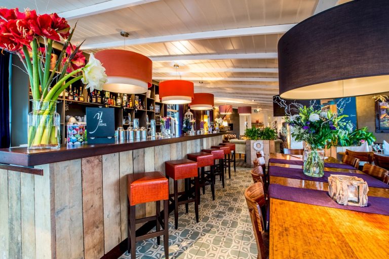 Stijlvol restaurantinterieur met een houten bar, oranje barkrukken, diverse bloemvazen en grote hanglampen.