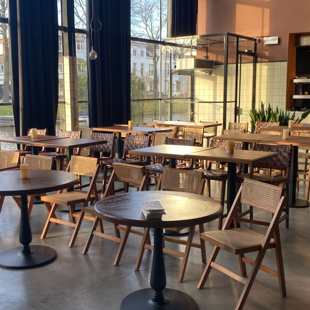 Sfeervol café interieur met houten tafels en rieten stoelen, badend in zonlicht, uitzicht op gracht.