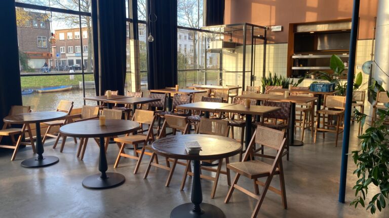 Sfeervol café interieur met houten tafels en rieten stoelen, badend in zonlicht, uitzicht op gracht.