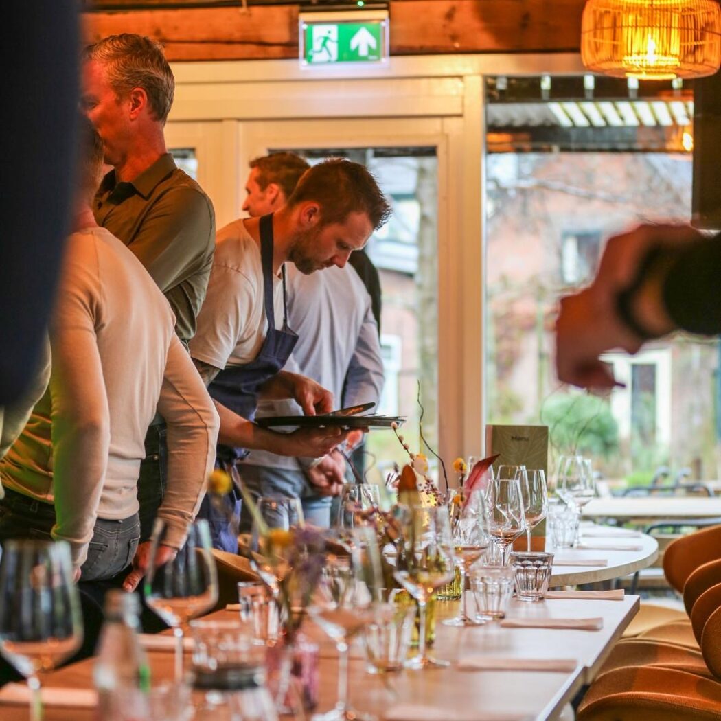 Medewerker in blauw schort kijkt naar een tablet bij de gedekte tafels in een warm verlicht restaurant.