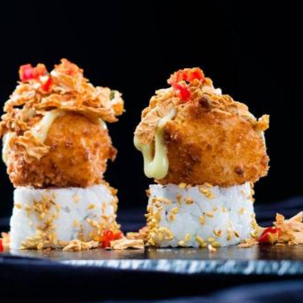 Twee makirollen met krokante gefrituurde topping, sesamzaadjes en druipende saus op een donkere plaat.