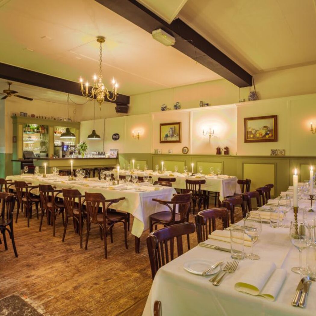 Ruime, sfeervolle eetzaal met lange, wit opgedekte tafels vol kaarsen, houten stoelen en een bar op de achtergrond.