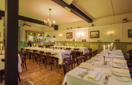 Ruime, sfeervolle eetzaal met lange, wit opgedekte tafels vol kaarsen, houten stoelen en een bar op de achtergrond.