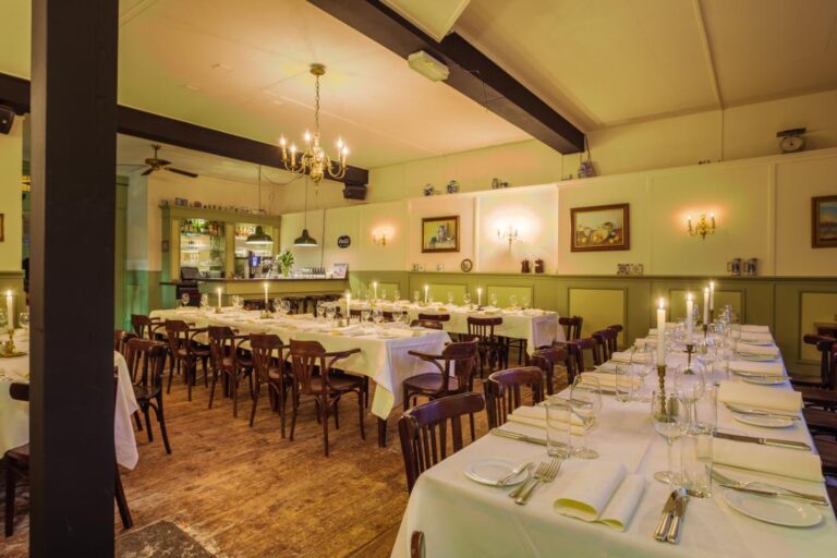 Ruime, sfeervolle eetzaal met lange, wit opgedekte tafels vol kaarsen, houten stoelen en een bar op de achtergrond.