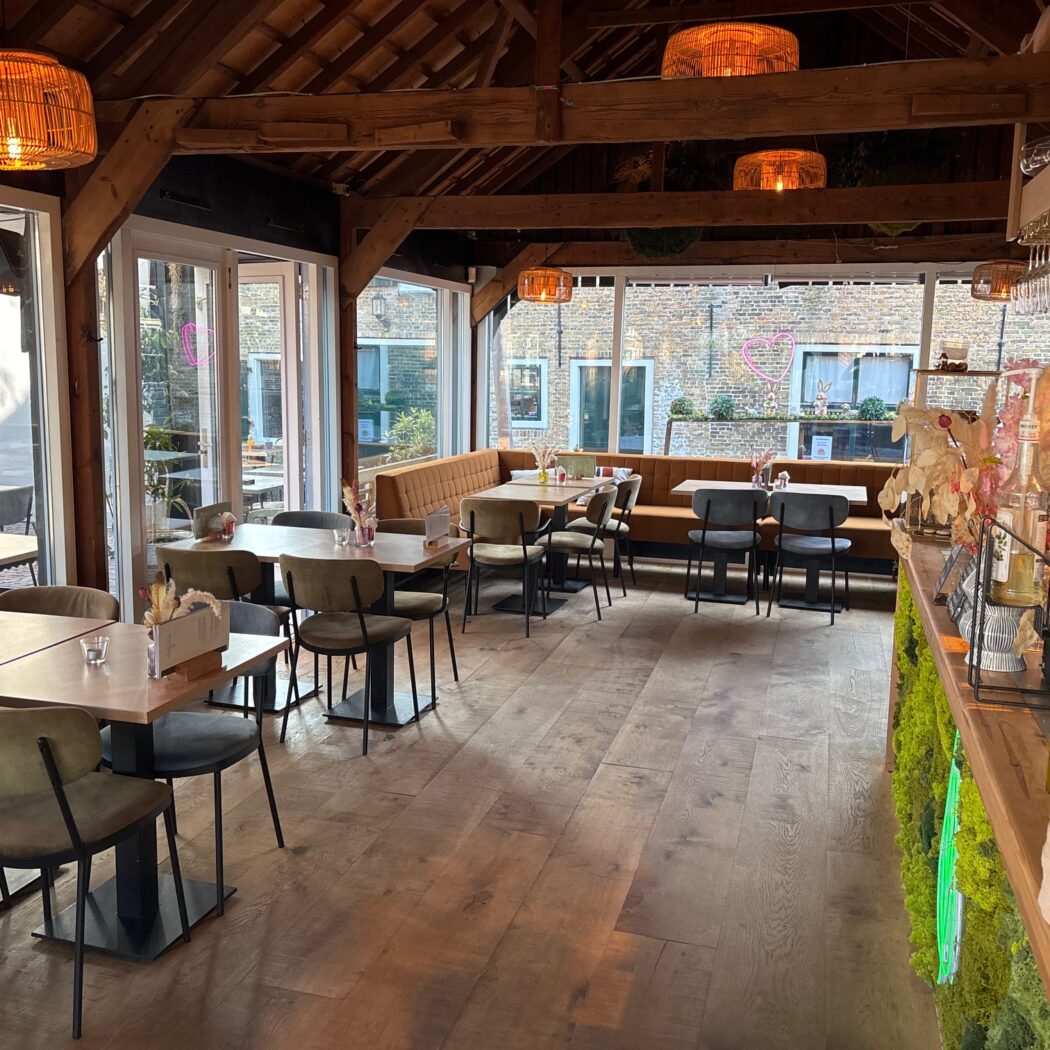 Warm café interieur met houten balkenplafond, rieten lampen, olijfgroene stoelen en een bar met groene moswand.