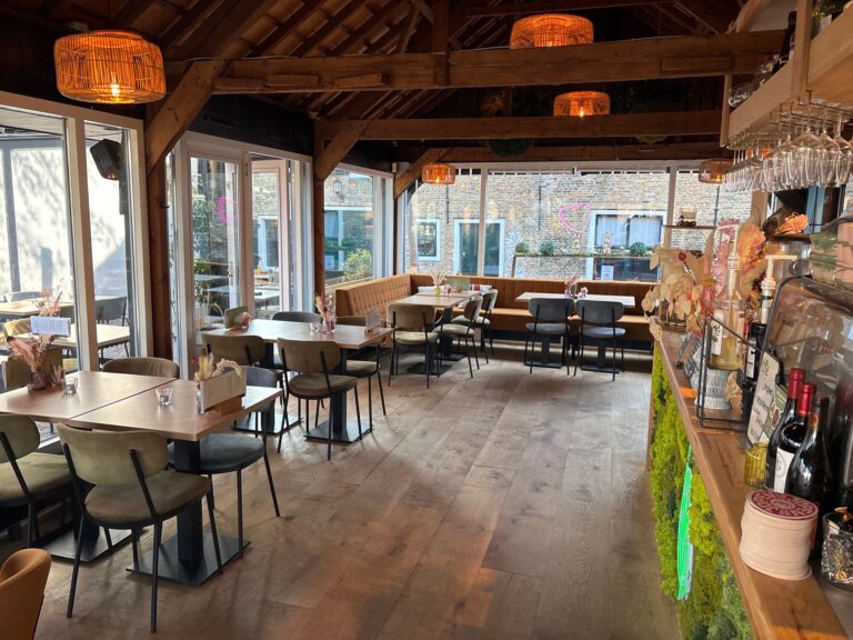 Warm café interieur met houten balkenplafond, rieten lampen, olijfgroene stoelen en een bar met groene moswand.