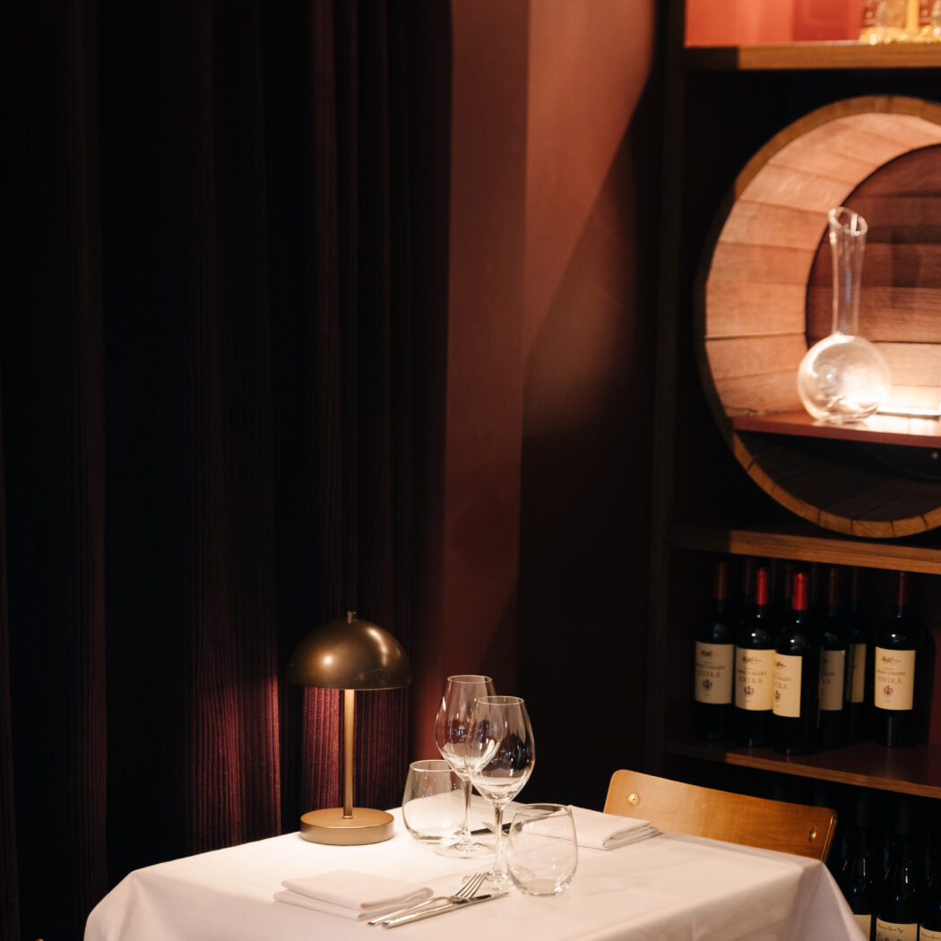 Gezellige restauranttafel met wit linnen, wijnglazen en een tafellamp, met rode gordijnen en wijnrekken.