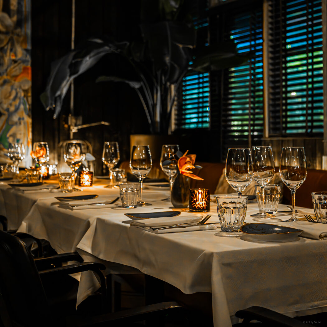 Sfeervol gedekte tafel met witte linnen, glazen, bestek, kaarsen en een orchidee in een restaurant.