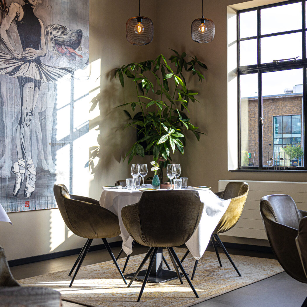 Zonnig restaurant met gedekte ronde tafels, olijfgroene fluwelen stoelen, grote plant en ballerina schilderij.
